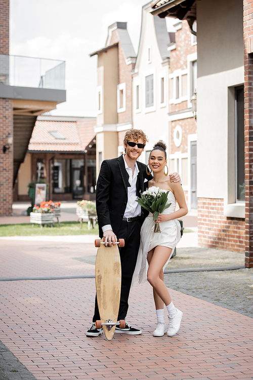 happy multiethnic couple with longboard and flowers looking at camera, wedding in european city