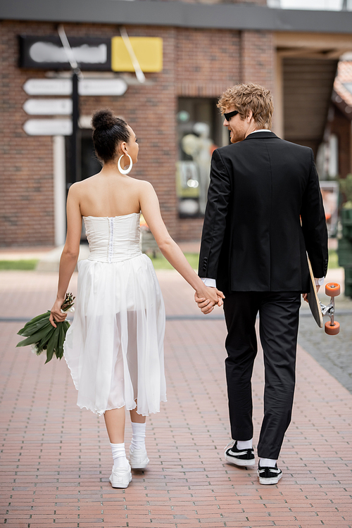 back view of multiethnic couple with longboard and flowers holding hands, walking in city, wedding