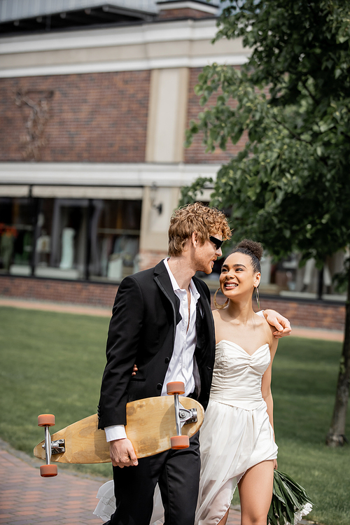 redhead groom with longboard walking with african american bride, wedding attire, sunglasses