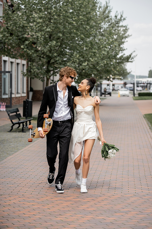 cheerful interracial newlyweds walking with longboard and flowers in european city