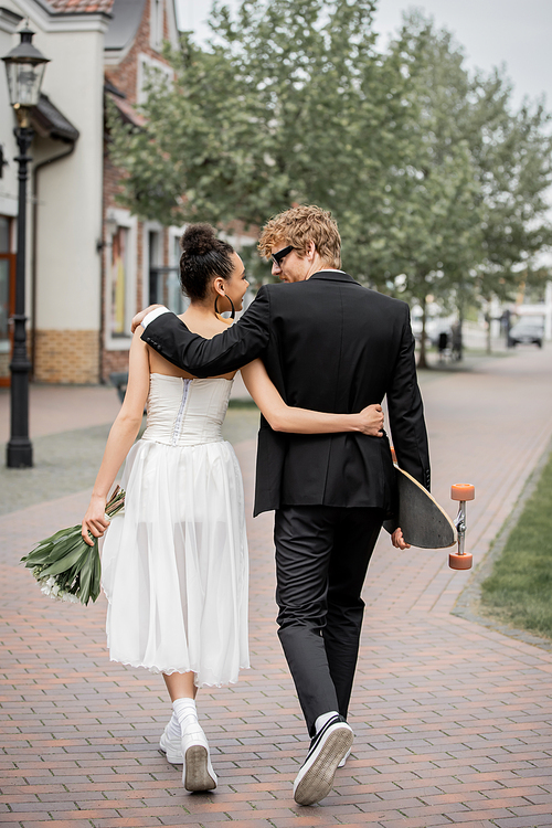 back view of stylish interracial newlyweds with flowers and longboard embracing and walking in city