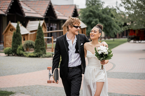 cheerful interracial couple walking in city with longboard and flowers, wedding attire, sunglasses