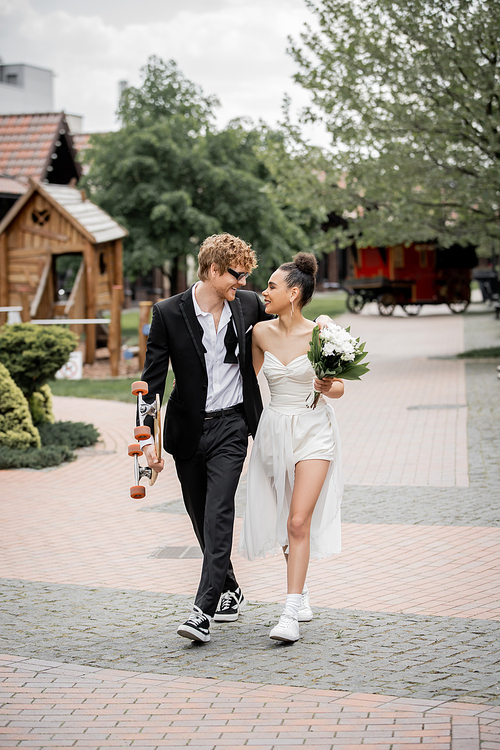 happy and stylish multiethnic couple walking with longboard and flowers, smiling at each other