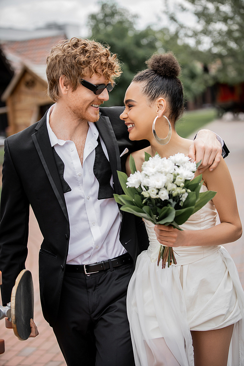 stylish smiling groom with longboard embracing african american bride with flowers on urban street