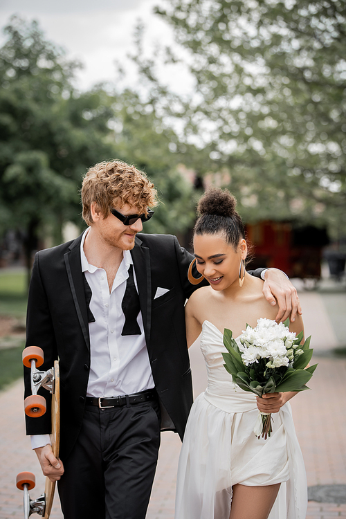 redhead groom in sunglasses embracing african american bride, walking with longboard and flowers