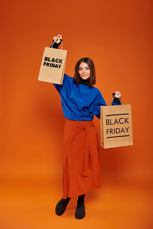 full length, pretty woman in autumn outfit holding shopping bags on orange backdrop, black friday