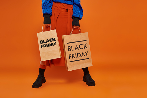 cropped woman in autumn skirt and boots holding shopping bags on orange backdrop, black friday