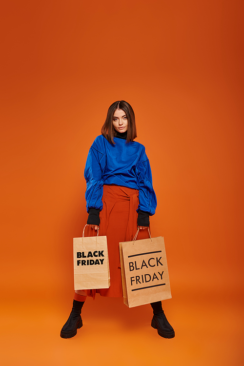 full length, brunette woman in autumn outfit holding shopping bags on orange backdrop, black friday