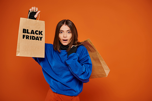 shocked woman holding shopping bags with black friday letters on orange backdrop, sales season