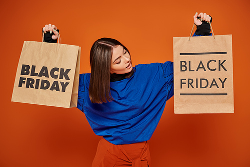 brunette woman holding shopping bags with black friday letters on orange backdrop, sales season