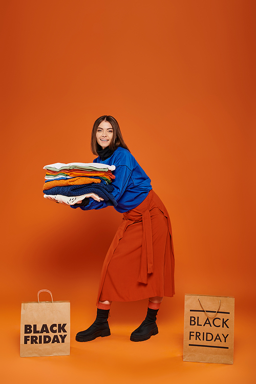 happy woman holding stack of autumnal clothes near shopping bags with black friday letters on orange