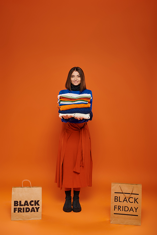 joyful woman holding stack of warm clothes near shopping bags with black friday letters on orange
