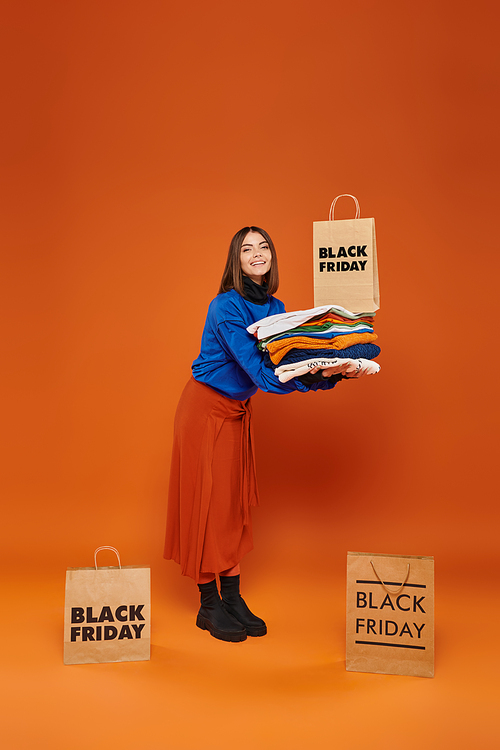 positive woman holding stack of warm clothes and shopping bag on orange backdrop, black friday