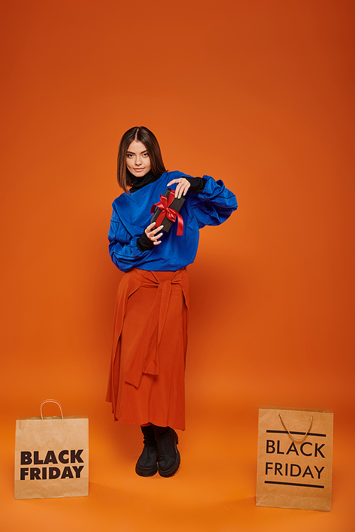 happy woman holding wrapped present near shopping bags on orange backdrop, black friday sales
