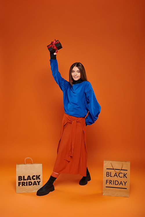 cheerful woman holding wrapped present near shopping bags on orange backdrop, black friday discounts
