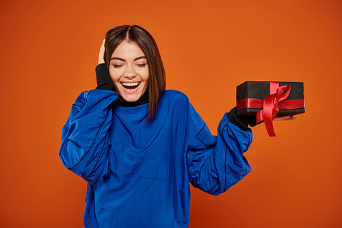 happy woman with brunette hair holding wrapped present on orange background, black friday