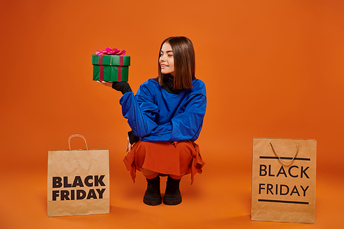 happy woman holding wrapped present and sitting near shopping bags on orange backdrop, black friday