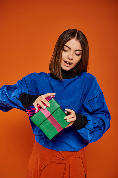 pretty woman with brunette hair opening wrapped present on orange background, Merry Christmas