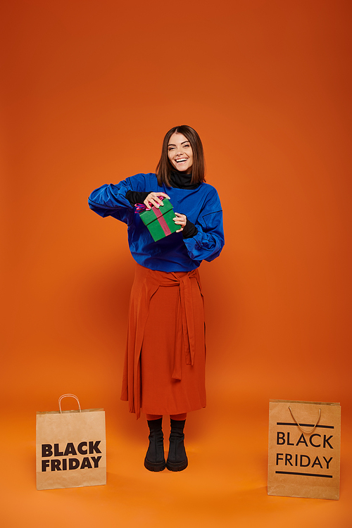 happy woman opening present near shopping bags with black friday letters on orange background