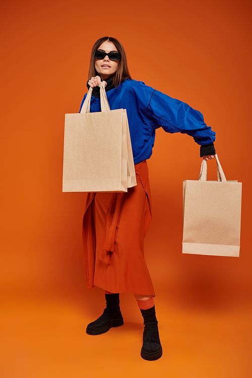brunette woman in stylish sunglasses holding shopping bags on orange backdrop, black friday sales