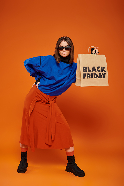 stylish woman in trendy sunglasses holding shopping bag with black friday letters on orange backdrop