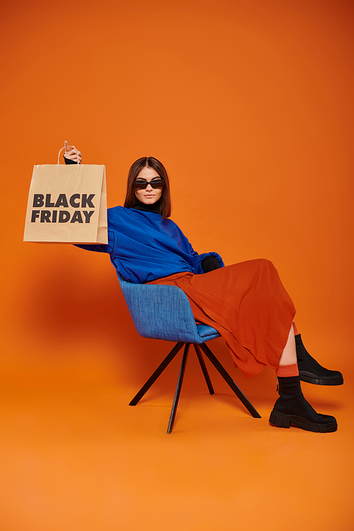 woman in sunglasses and autumnal attire holding black friday shopping bag and sitting on armchair