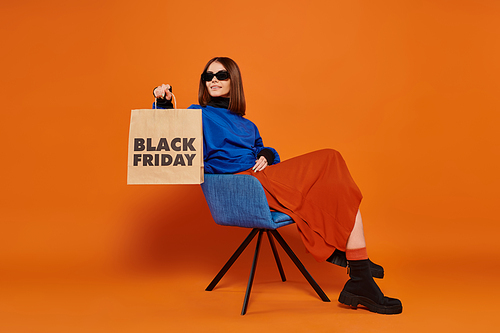 happy woman in stylish sunglasses holding black friday shopping bag and sitting on armchair