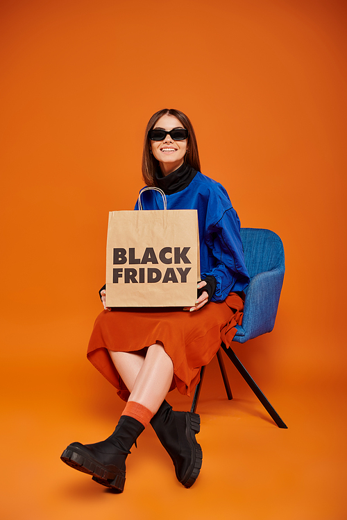 cheerful woman in stylish sunglasses holding black friday shopping bag and sitting on armchair