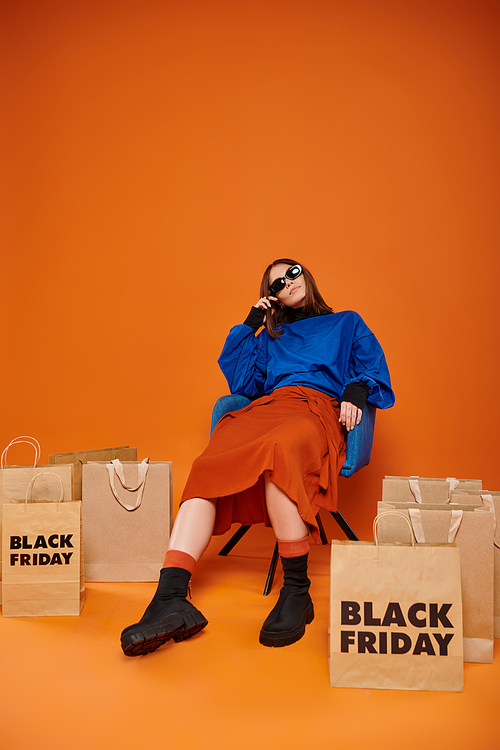 tired woman in stylish sunglasses sitting on armchair near black friday shopping bags on orange