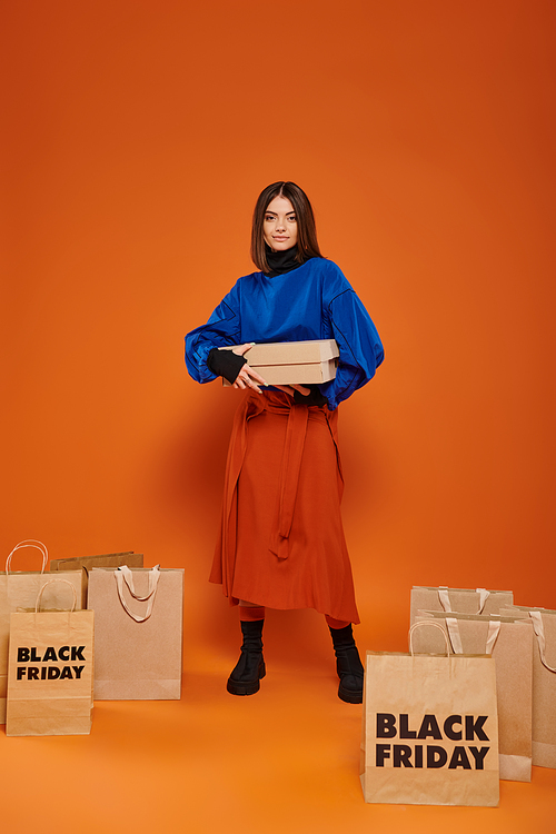 happy woman holding carton box and standing near shopping bags with black friday letters, orange