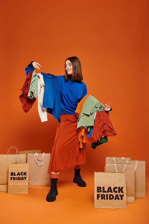 happy woman with brunette hair holding colorful autumnal clothes,  near black friday shopping bags
