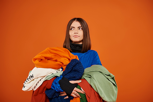 pensive young woman with brunette hair holding pile of colorful autumnal clothes, black friday