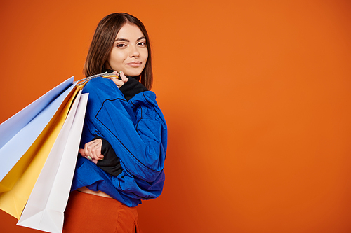 positive woman in stylish autumnal clothes holding shopping bags on black friday, orange backdrop