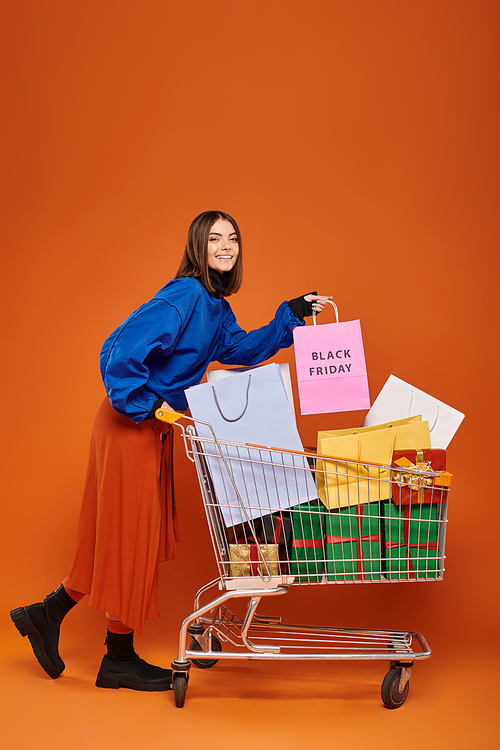 joyful woman standing with cart full of shopping bags with black friday letters on orange backdrop