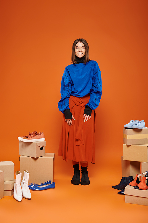 happy woman in autumnal clothes standing near boxes with different shoes on orange, black friday