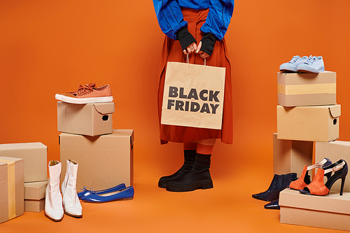 cropped woman in autumnal clothes standing near boxes with different shoes on orange, black friday