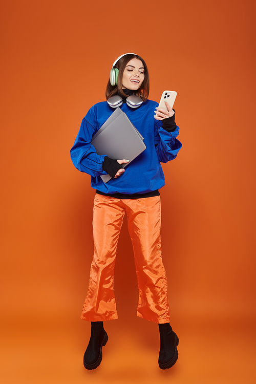 happy woman in headphones and autumnal outfit using smartphone and holding laptop, cyber monday