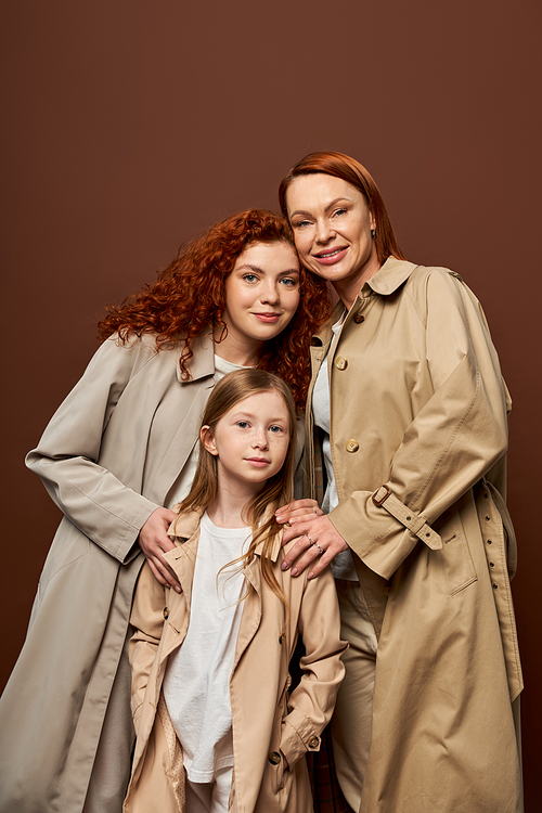 three generations, cheerful women and girl posing in autumn trench coats on grey background, fashion