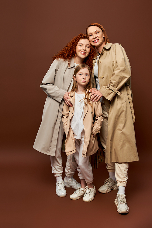 three generations, happy redhead family posing in autumn trench coats on grey background, fashion