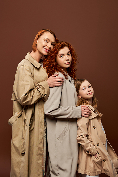 three generations, happy redhead women and cute girl in trench coats posing on brown background