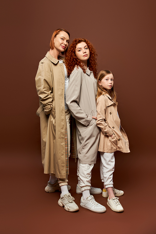 three generations, redhead women and girl posing in coats on brown backdrop, autumnal attire