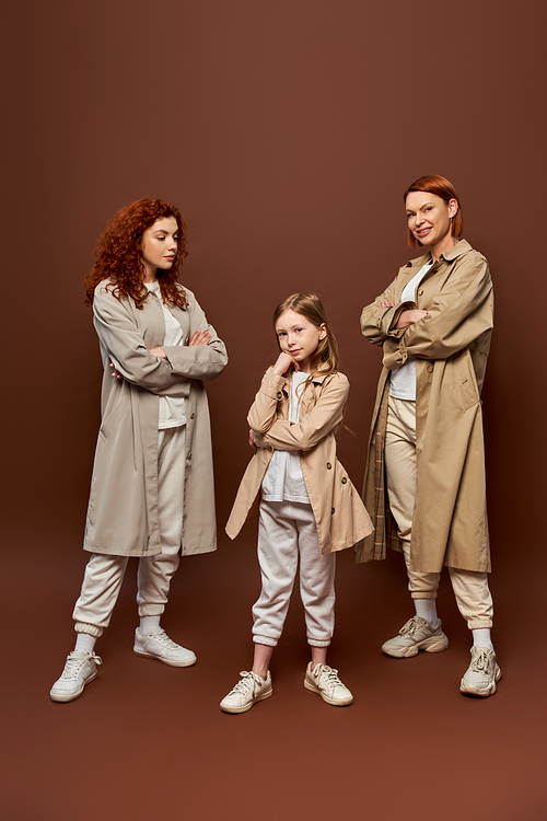 three generations, women in coats posing and looking at girl on brown backdrop, redhead family