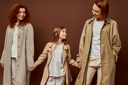 three generations, cheerful redhead family in coats holding hands on brown backdrop, women and girl
