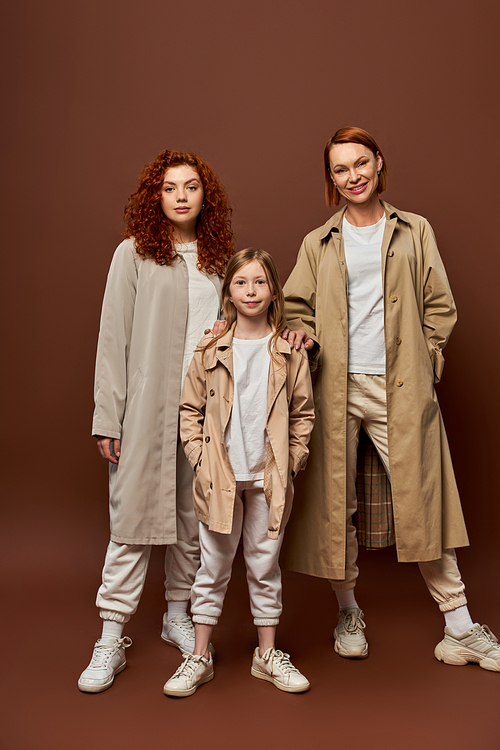female generation, redhead family standing together in outerwear on brown backdrop, women and girl