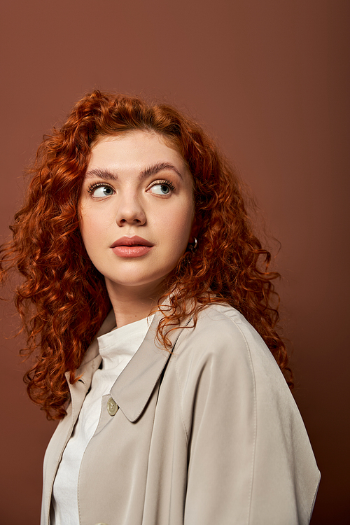 portrait of dreamy redhead woman with curly hair looking away on brown background, autumn colors