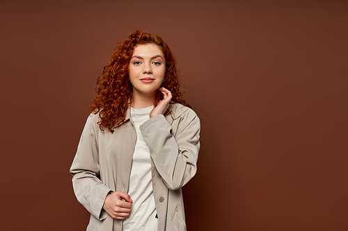 portrait of pleased woman with red curly hair looking at camera on brown background, autumn colors