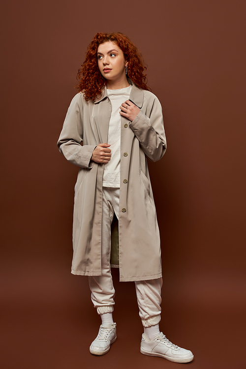 young woman with red curly hair posing in autumn coat and joggers on brown background, fall season