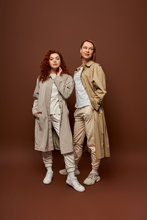 two generations, redhead women in trendy autumn clothes posing on brown background, trench coats