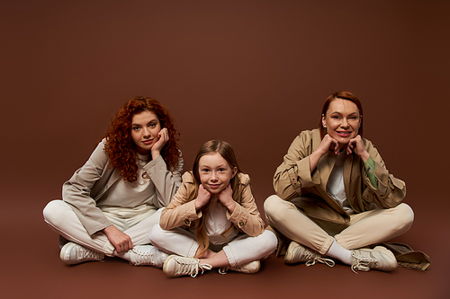 three generations of women looking at camera and sitting in coats on brown backdrop, cute girl