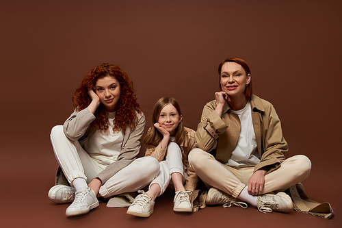 happy redhead family of three female generations sitting on brown background, autumn coats